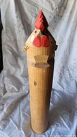 Front view of handmade wooden rooster figurine showing its carved facial features, red painted comb and wattles, cylindrical body with natural wood stain.