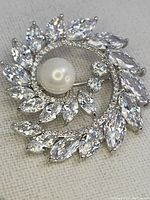 Close-up photo of the brooch showing the arrangement of marquise-cut clear stones and a pearl center, highlighting the prong settings and silver-tone metal backing.