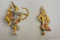 Front view of two antique figural brooches on fabric background showing detailed gold and silver tones, one figure holding bow and arrow, other standing pose
