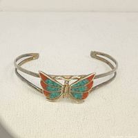 Metal cuff bracelet with butterfly ornament adorned with turquoise and coral chip inlay.
