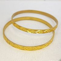 Two antique gold-colored bangles displayed side by side on white fabric, one engraved with floral patterns.