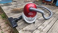 Samsung Easy 1300 vacuum cleaner shown from the side on wooden surface, displaying the hose and floor brush attachment