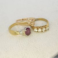 Three vintage rings displayed on fabric background. Includes one garnet ring, one with four clear stones, and one delicate rose gold-tone ring with three small stones.