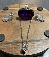 Top view of the ring with large oval purple faux amethyst cabochon, rhinestone wheel pendant necklace chain laid down, and tristar rhinestone dangling earrings on dark wooden display