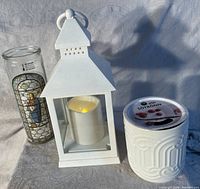 Photo showing three items: tall religious candle in glass, white battery lantern with flameless candle inside, and sealed Ikea scented candle in ceramic holder.