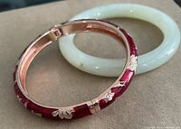 Two bangles shown together on a surface, one red enamel hinged bangle with floral decoration and one pale green jadite bangle.