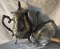 Tea/coffee pot standing upright next to ornate tray against white fabric backdrop.