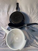 Two black Lagosina Renaissance non-stick pans and one white Rubbermaid plastic strainer, shown stacked with handles visible.