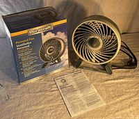 Duracraft personal fan shown outside the box with instruction manual, placed on a fabric surface under warm lighting.