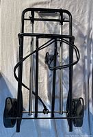 Folded black metal frame dolly with large black rubber wheels and black bungee cord wrapped around the frame.