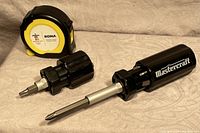 Two Mastercraft multi-change screwdrivers and a small RONA tape measure arranged on a light fabric surface.