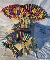 Hawaiian-themed party favors including floral fabric fans, wooden maraca shaker pens with colorful patterns, packaged beaded attachments, and various wristbands displayed on a light fabric surface.