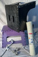 Image showing black and purple foldable step stools stacked with a roll of the Rubbermaid bath mat, white over-the-cabinet hook, and three metallic cabinet knobs laid in front.