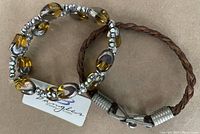 Two bracelets on neutral background: amber-topaz rhinestone stretch bracelet with silver accents and attached designer tag, laid next to brown leather braided bracelet with pewter clasp showing wear.