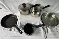 Set of five pots and pans arranged on white cloth with various sizes and handles, showing condition.
