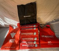 Photo showing 6 reusable shopping bags; four red Shoppers Drug Mart bags stacked horizontally, one red Canada's Store bag on the left, one red Staples bag on the right and one black Rogers bag standing upright at the back.