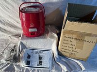 Red Perfect Cooker rice cooker with power cord, spoon, manual, and recipe book all laid out on a white surface with the original box.