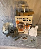 Photo showing the assembled parts of the chopper including the transparent 3 cup bowl, SmartChop attachment, lid, and manuals, laid out on a cloth background.