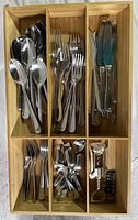 Top-down view of stainless steel spoons, forks, and knives arranged neatly in wooden utensil case compartmentalized into six sections.