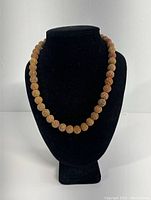 Front view of carved stone necklace displayed on black bust form showing 20 carved beads in light beige and reddish tones.
