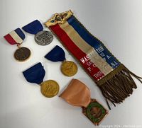 Top-down photo showing six commemorative medals laid out alongside a colorful fabric ribbon with gold fringe and ornate metal top piece. Medals vary in shape, color, and ribbon style.