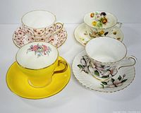 Full view of four teacups and matching saucers showing varied floral and patterned designs including red floral, yellow with flower interior, multicolored flowers, and purple flowers with gold trim.