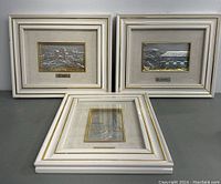 Three framed embossed metal pictures stacked on a table, showing white frames with gold detailing and artist nameplates under the embossed artwork.