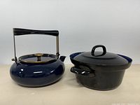 Front angle view of the dark blue enamelled metal kettle with brass-colored lid handle and black handle, and the black lidded pot showing the handle and shape.