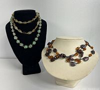 Three beaded necklaces displayed on two bust stands: one black and one white. Greenish stone bead necklace on black bust; brown amber and smoky bead necklace on white bust; third brown beaded necklace draped on black bust.