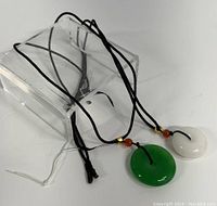 Photo of two jade pendant necklaces laid flat showing green and white round pendants on black braided ropes with amber and gold beads.