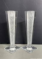 Two tall clear crystal candle holders with pentagonal bases and etched leaf motifs displayed against a black background