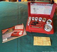Opened red plastic Milwaukee carrying case displaying 12 bi-metal hole saws of different sizes, 3 arbors with pilot drills (one pilot drill missing) and original paperwork/manuals alongside the kit box.