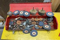Red metal toolbox open showing many assorted grinding stones and grinding wheels inside and on top