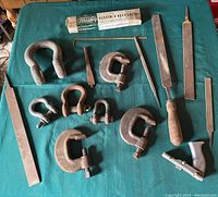 Photo of assorted vintage iron C-clamps, small shackles with screw pins, various metal files, file holder, and packaged flexible belt lacing on a green table.