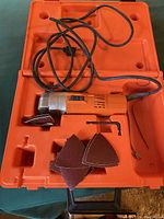 Fein electric triangle detail sander inside orange carrying case with power cord and sanding pads.