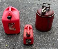 Photo showing all three gas cans: large 5 gallon red plastic can, smaller 1 gallon red plastic can, and vintage red metal 5 gallon can with handle and spout on asphalt ground.
