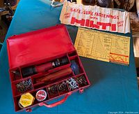 Open metal case showing piston drive tool inside, assorted fasteners, cartridges, cloth gloves, and a selector chart attached to the lid.