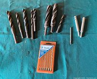 Loose large drill bits, small adjustable chuck, smaller drill bit, and packaged David Universal drill bit set on green cloth backdrop.