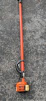 Full length view of orange pole pruner with engine and handle, placed on ground.