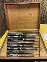 Wooden box containing 8 adjustable steel reamers arranged in slots.