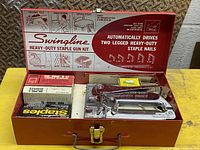 Opened red metal case showing Swingline staple gun on right and several boxes of staples on left; printed instructions on inside lid.
