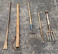 Full view of all five vintage garden tools on pavement showing crow bar, pitch forks, dandelion weeder, and log roller
