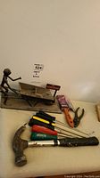 Photo showing the handmade bolt sculpture of a man pushing a wheelbarrow alongside various hand tools including claw hammer, screwdrivers, pliers, and paint scraper.