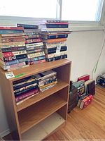 Books stacked on a small wooden bookcase and on the floor beside it near a window