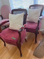 Pair of vintage wooden chairs with cane backrests and maroon velvet cushioned seats, with white pillows on each chair, positioned on wood flooring in front of a window.