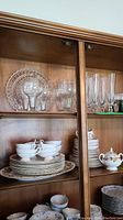 Cabinet shelf with stacked plates, bowls, tea cups, and various glassware including wine glasses and tumblers.