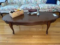 Wooden coffee table with scalloped edges, curved legs, medium to dark brown finish, placed on hardwood floor with floral upholstered sofa in background. Items on table not included.