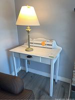 White painted wood desk with single drawer and table lamp sitting on top, lamp turned on, in a corner near a striped chair.