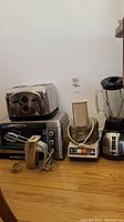 Full view of all kitchen appliances on wooden floor against wall including Black & Decker blender, GE food processor, four slice toaster, toaster oven, and vintage Sunbeam Mixmaster hand mixer.