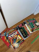 Row of Marvel and DC comic magazines on wood floor, shows colorful covers with characters including Spider-Man and Thor.